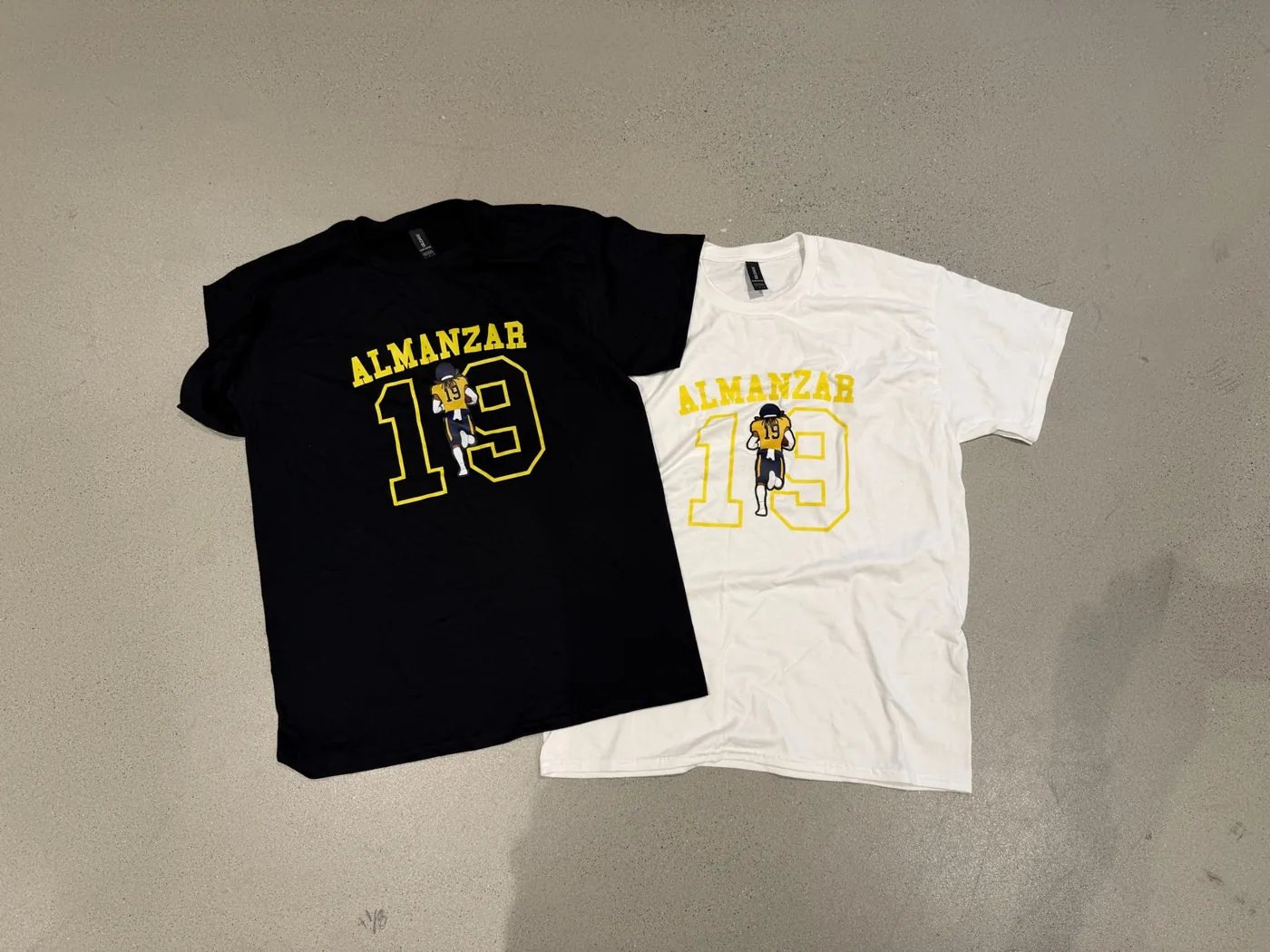 A black and a white Almanzar 19 football tee laid side by side on a concrete floor